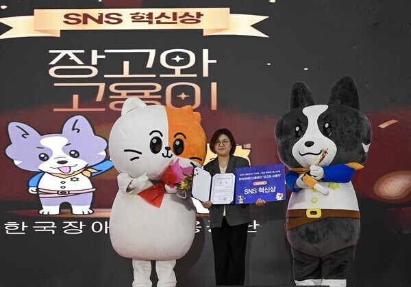 “SNS 혁신상 시상식 현장 사진. 무대 중앙에서 한 여성이 상장과 ‘SNS 혁신상’이라고 적힌 상패를 들고 서 있으며, 양옆에는 캐릭터 인형 탈을 쓴 두 마스코트가 함께 서 있다. 무대 뒤 대형 화면에는 ‘SNS 혁신상’과 ‘잡고와 고용이’라는 문구가 표시되어 있다. 수상 축하를 기념하는 공식 시상 장면이다.”  원하면